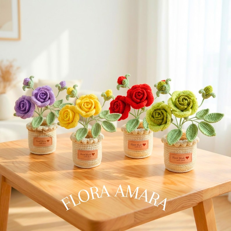 May include: Four crocheted rose arrangements in small, cream-coloured woven pots. Each pot features a brown label with the text Hand Made with a heart icon. The roses are purple, yellow, red, and green, each with green crocheted leaves and stems.