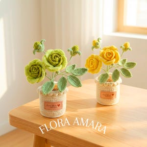 May include: Two crocheted potted plants featuring green and yellow roses with buds and leaves. Each plant sits in a cream-coloured crocheted pot with a tan leather-like label that reads Hand Made with love and includes a small red heart icon.
