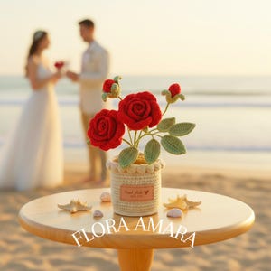 May include: A crocheted red rose arrangement in a cream-coloured pot with a scalloped rim. The pot features a brown rectangular tag with the text "Hand Made with care" and a heart icon. The text "FLORA AMARA" is printed at the bottom of the image.