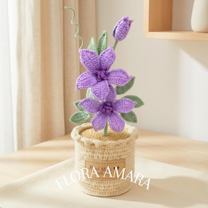 May include: A handmade crochet flower arrangement with two purple flowers, a bud, and green leaves in a beige woven pot. The pot has a label with the text "Hand Made with love" and the brand name "FLORA AMARA".