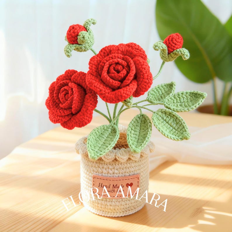 May include: A crocheted flower arrangement featuring two fully bloomed red roses and two red rosebuds with light green leaves and stems. The flowers are set in a cream-coloured crocheted pot with a peach-coloured label reading Hand Made with love FLORA AMARA.