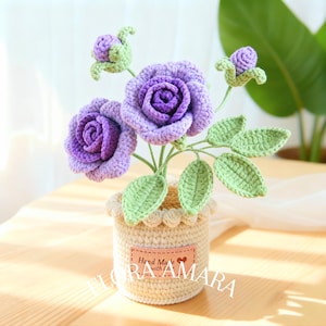May include: A crocheted arrangement featuring two purple roses and two small buds with light green leaves, set in a cream-coloured crocheted pot. A rectangular tan label on the pot displays the text Hand Made with love and FLORA AMARA.
