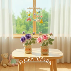 May include: Two crochet flower arrangements in beige pots with the text FLORA AMARA. One pot contains purple roses with green leaves, and the other contains pink roses with green leaves. Each pot features a small rectangular label with text.