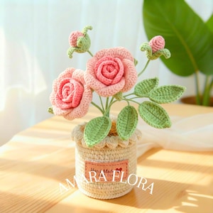 May include: A crochet rose arrangement featuring two blooming pink roses and two small rosebuds with green leaves. The flowers are set in a cream-coloured crocheted pot with a rectangular leather-like tag that reads Hand Made with love AMARA FLORA.
