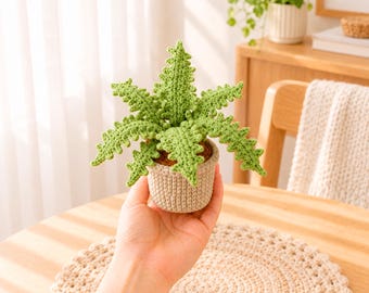 May include: A crocheted fern plant with bright green, serrated leaves potted in a small, beige, cylindrical crocheted container. The plant features multiple textured fronds radiating outward from a brown, soil-like center base.