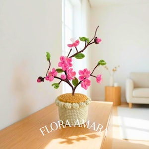 May include: A handmade crochet floral arrangement with pink flowers, green leaves, and brown stems. The flowers are in a yellow and cream-coloured crochet pot. The text "FLORA AMARA" is visible on the front of the pot.