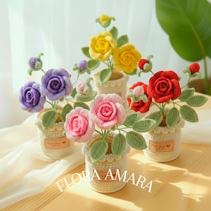 May include: A selection of handcrafted crochet roses in shades of purple, pink, yellow, and red. Each rose is presented in a small, cream-coloured crocheted pot with green leaves. The text "FLORA AMARA" is displayed.
