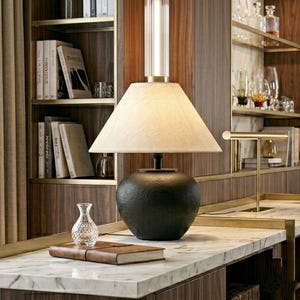 Puede incluir: Una lámpara de mesa negra con una base texturizada y una pantalla de color crema. La lámpara está sobre una encimera de mármol, con un pequeño jarrón de cristal y un libro encuadernado en cuero cerca. El fondo presenta una estantería de madera.