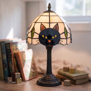 Könnte beinhalten: Dekorative Tischlampe mit einem Lampenschirm aus Buntglas mit einem schwarzen Katzendesign. Der Schirm hat einen cremefarbenen Hintergrund mit schwarzen Konturen und grünen Blattakzenten. Der Sockel ist dunkelbraun mit einem detaillierten Muster.