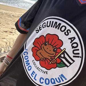 K&ouml;nnte beinhalten: Schwarzes Mesh-Trikot mit einem runden Grafikdesign, das einen Frosch in einer roten Blume zeigt, der eine Flagge h&auml;lt. Der Text um die Grafik lautet "SEGUIMOS AQUI" und "COMO EL COQUI". Das Trikot hat blaue und rote Streifen an den &Auml;rmeln.
