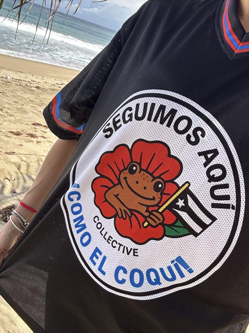 K&ouml;nnte beinhalten: Schwarzes Mesh-Trikot mit einem runden Grafikdesign, das einen Frosch in einer roten Blume zeigt, der eine Flagge h&auml;lt. Der Text um die Grafik lautet "SEGUIMOS AQUI" und "COMO EL COQUI". Das Trikot hat blaue und rote Streifen an den &Auml;rmeln.