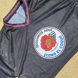K&ouml;nnte beinhalten: Schwarzes Sporttrikot mit einem runden Emblem, das einen Frosch mit einer Flagge in einer roten Blume zeigt. Das Emblem tr&auml;gt den wei&szlig;en Text "SEGUIMOS AQUI" und "COMO EL COQUI!". Das Trikot hat blaue und rote Verzierungen.