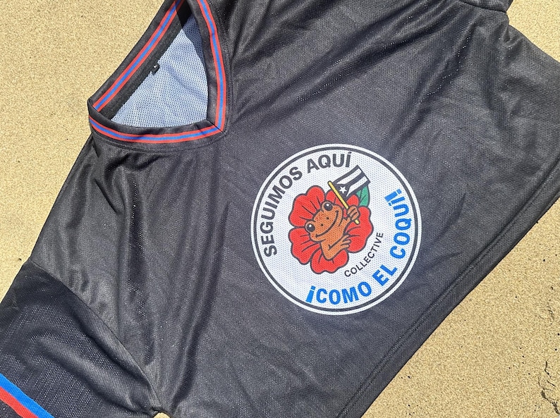 K&ouml;nnte beinhalten: Schwarzes Sporttrikot mit einem runden Emblem, das einen Frosch mit einer Flagge in einer roten Blume zeigt. Das Emblem tr&auml;gt den wei&szlig;en Text "SEGUIMOS AQUI" und "COMO EL COQUI!". Das Trikot hat blaue und rote Verzierungen.