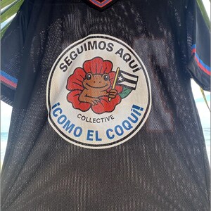 K&ouml;nnte beinhalten: Schwarzes Mesh-Trikot mit V-Ausschnitt und kurzen &Auml;rmeln. Das Trikot hat eine kreisf&ouml;rmige Grafik mit einem Frosch, der eine Flagge h&auml;lt, und den Text "SEGUIMOS AQUI" und "COMO EL COQUI!" in Wei&szlig; und Blau.