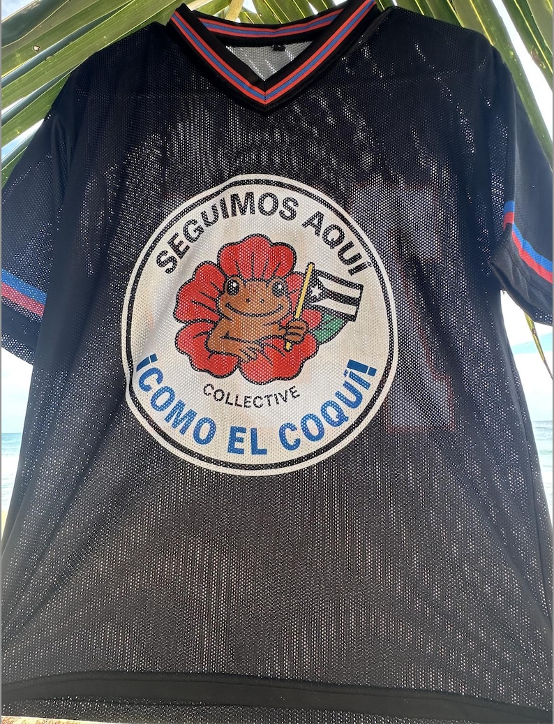 K&ouml;nnte beinhalten: Schwarzes Mesh-Trikot mit V-Ausschnitt und kurzen &Auml;rmeln. Das Trikot hat eine kreisf&ouml;rmige Grafik mit einem Frosch, der eine Flagge h&auml;lt, und den Text "SEGUIMOS AQUI" und "COMO EL COQUI!" in Wei&szlig; und Blau.