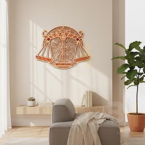 May include: Wooden Libra zodiac sign wall art. The layered design features the scales of justice, a star, and intricate geometric patterns. The artwork is made of light and dark brown wood, creating a 3D effect. It is mounted on a neutral-colored wall.