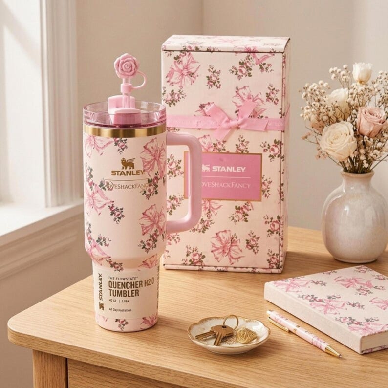 May include: A pink floral Stanley Quencher H2.0 tumbler with a handle, a matching gift box, and a small dish with keys on a wooden table. The tumbler has a floral design and the text "The Flowstate Quencher H2.0 Tumbler 40 oz".