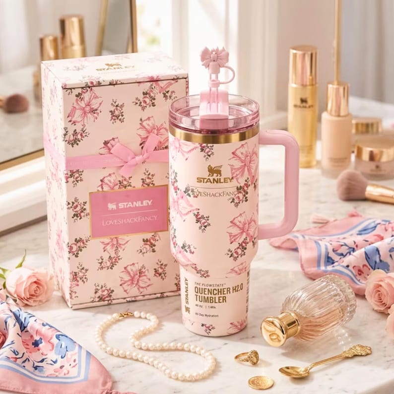 May include: A pink Stanley Quencher H2.0 Tumbler with a floral and bow design, a matching gift box, and a pink handle. The tumbler has a capacity of 40 oz. and features the text "LOVESHACKFANCY".