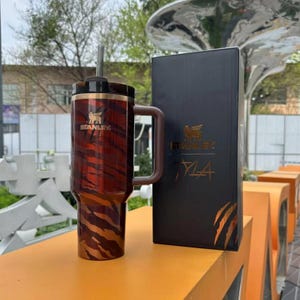 K&ouml;nnte beinhalten: Ein gro&szlig;er, isolierter Becher mit einem Tigerstreifenmuster in Rot- und Braunt&ouml;nen, mit Griff und Strohhalm. Der Becher ist mit dem Logo "Stanley" versehen. Eine schwarze Box mit goldfarbenen Buchstaben und Krallenmarkierungen befindet sich neben dem Becher.