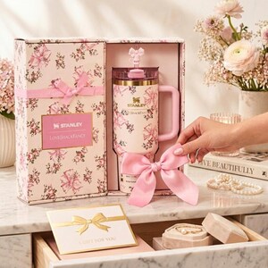 May include: A pink floral-patterned Stanley tumbler with a matching gift box. The tumbler has a handle and a straw. The gift box is tied with a pink ribbon. The text "Stanley LovesShackFancy" is visible on the box.