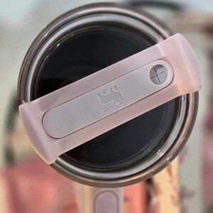 May include: Close-up of a pink and gray Stanley tumbler lid. The lid features a sliding closure with the Stanley logo and a cross-shaped detail. The tumbler's interior is dark, and the lid is angled across the opening.