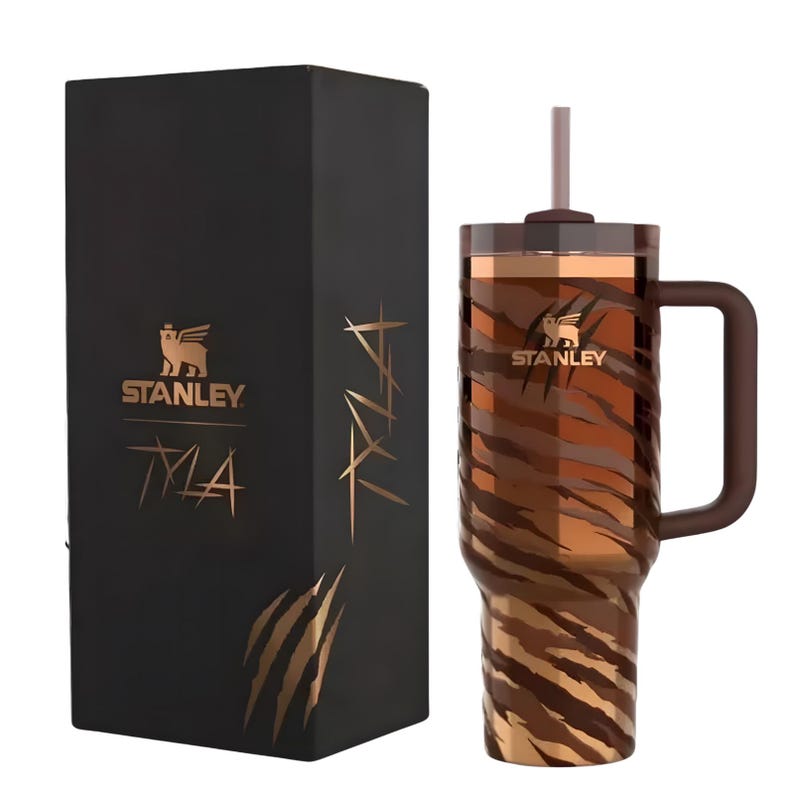 K&ouml;nnte beinhalten: Ein isolierter Stanley-Becher mit braunem Tigerstreifenmuster, einem Griff und einem Strohhalm. Der Becher steht neben einer schwarzen Box mit dem Stanley-Logo und dem Text "TLA" in Gold.
