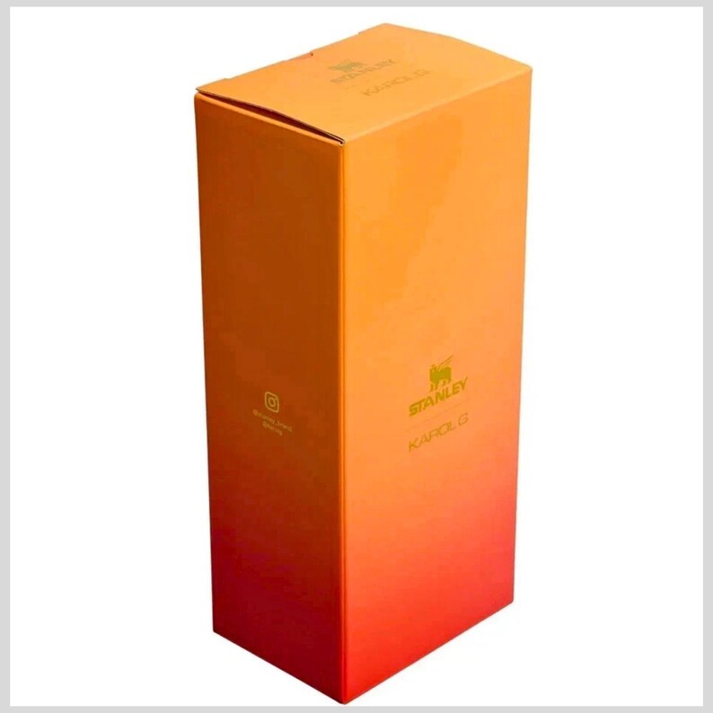 May include: A rectangular cardboard product box featuring a vertical gradient color scheme that transitions from orange at the top to red at the bottom. The box displays the Stanley logo and the text KAROL G in gold lettering on the front face.