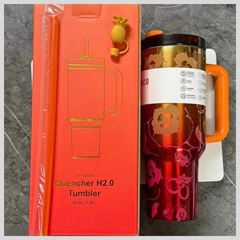 May include: A red and gold floral patterned 40 oz tumbler with an orange handle and lid, accompanied by a matching orange straw and a small pineapple-shaped charm. The product is displayed next to its original red packaging box with text.