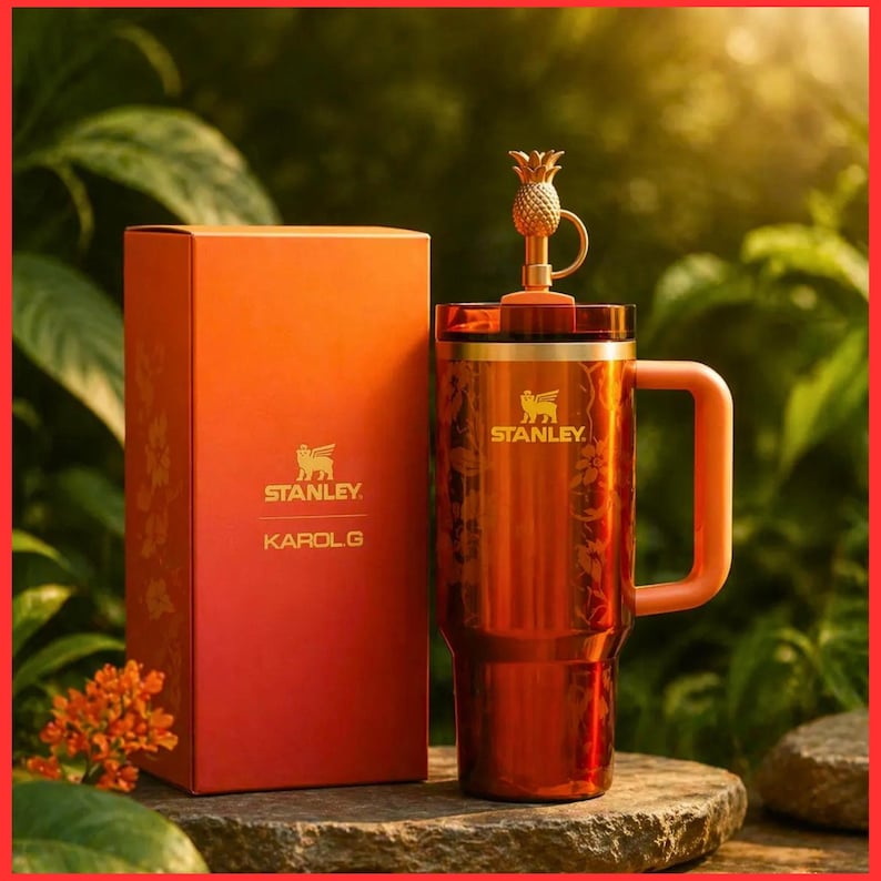 May include: A metallic orange Stanley tumbler with a matching handle and a gold pineapple-shaped topper on the lid. The tumbler features a floral pattern and the Stanley logo. Beside it stands an orange box labeled Stanley and Karol G.