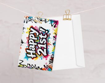 May include: A white greeting card with the text "Happy 21st!" in a graffiti style, featuring vibrant colors like pink, yellow, and blue. The card is clipped to a white envelope with gold paper clips, hanging against a neutral background.