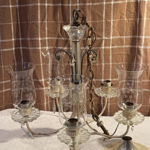 May include: An antique chandelier with five arms, featuring clear glass shades etched with a delicate pattern. The fixture has a silver finish with a decorative chain and a scalloped base. The shades are shaped like tulip glasses.