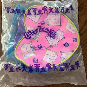 May include: A heart-shaped Cabbage Patch Kids Sweetheart Purse by Avon, enclosed in clear plastic packaging. The purse features a bright pink surface with a repeating pattern of doll faces, hearts, and the Cabbage Patch Kids logo in purple.