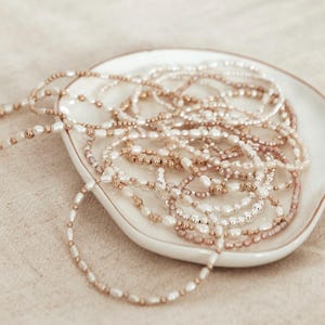 May include: A collection of delicate pearl and beaded necklaces in shades of white, gold, and rose gold, displayed on a white, scalloped-edge dish. The necklaces vary in design, with small, round beads and elongated pearls.