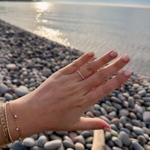 May include: A hand wearing a delicate ring and multiple gold bracelets with pearls. The ring features a series of small, evenly spaced stones. The bracelets are layered, with varying designs including small beads and delicate chains. The background shows a pebble beach and the ocean.