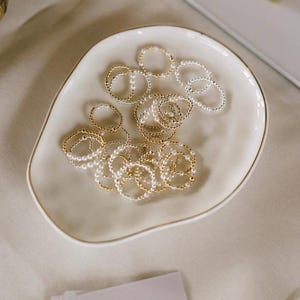 May include: A collection of beaded rings in gold and silver tones, displayed in a cream-colored, oval-shaped dish with a gold rim. The rings are various sizes and styles, with some featuring small pearl-like beads.