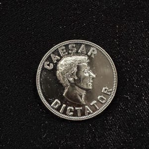 Könnte beinhalten: Eine silberne Münze mit dem Profil eines Mannes und den Worten "CAESAR DICTATOR" am Rand. Die Münze befindet sich vor einem schwarzen Hintergrund.