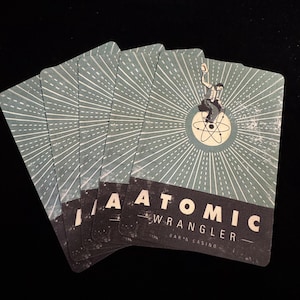 May include: A fanned deck of playing cards with a teal and white sunburst design. Each card features the text "ATOMIC WRANGLER BAR & CASINO" and an illustration of a man on an atom. The cards have a vintage aesthetic.