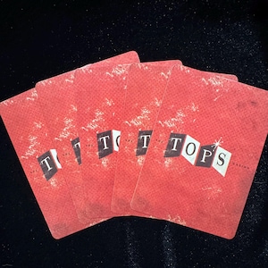 May include: A fan of five red playing cards with the word "TOPS" in white letters. The cards have a distressed, vintage look, with a textured background. The word "CASINO" is visible on the rightmost card.