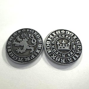 May include: Two silver-colored coins with detailed engravings. One coin features a lion rampant, the other a crown. Both coins have circular designs with text around the edges, suggesting they are historical or commemorative.