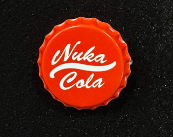 May include: A red bottle cap with the words "Nuka Cola" in white script. The cap has a ridged edge and is set against a black, textured background. The design is reminiscent of a popular video game.