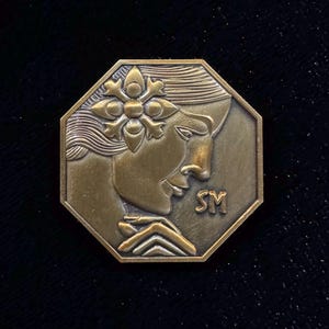 Könnte beinhalten: Achtseitige Münze in Bronzefarbe mit dem Profil einer Frau mit floralem Kopfschmuck. Die Münze hat die Initialen "SM" und ist vor einem dunklen Hintergrund platziert. Das Design ist detailliert und künstlerisch.