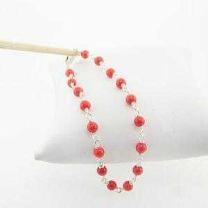 Puede incluir: Un brazalete de cuentas de coral rojo con una cadena de plata. El brazalete está hecho de pequeñas cuentas redondas y mide aproximadamente 18 cm de largo.