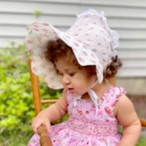 May include: A young child wearing a white bonnet with a floral pattern and a pink floral dress. The bonnet has a wide brim and a ruffle trim.