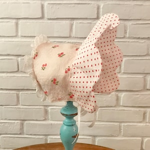May include: A white bonnet with a pink cherry print and a white polka dot trim. The bonnet has a white lace trim and a white ribbon tie.