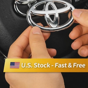 May include: A chrome Toyota emblem is held in front of a steering wheel. The emblem is oval-shaped with the Toyota logo in the center. The text "U.S. Stock - Fast & Free" is displayed on a yellow banner.