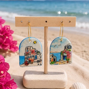 Può includere: Un paio di orecchini a forma di arco con scene costiere mediterranee dipinte a mano che raffigurano edifici bianchi, acqua blu e fiori rosa. Gli orecchini pendono da sottili cerchi in metallo dorato su un espositore in legno chiaro.