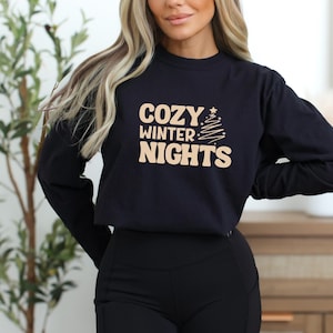 Könnte beinhalten: Schwarzes Langarmshirt mit dem Text "COZY WINTER NIGHTS" in Beige und einer Weihnachtsbaumgrafik. Das Shirt ist aus einem weichen Material gefertigt, ideal für Winternächte.