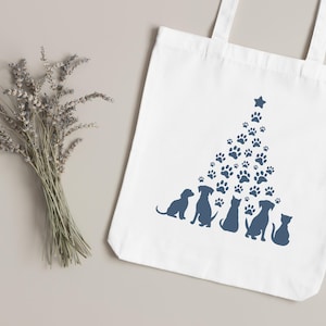May include: White tote bag with a Christmas tree design made of blue paw prints and a star. Silhouettes of dogs and a cat sit below the tree. A bundle of dried lavender is to the left.