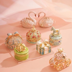 May include: An assortment of decorative trinket boxes in various shapes and designs. The collection includes swan figurines, crown-topped boxes, a rocking horse box, a gift box, and a carriage, all in pastel colors with ornate details.