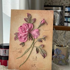 May include: A textured artwork featuring a pink rose with green stems and leaves against a beige background. The rose is in full bloom with detailed petals. The artwork has a vintage aesthetic, with script-like text and a signature at the bottom.